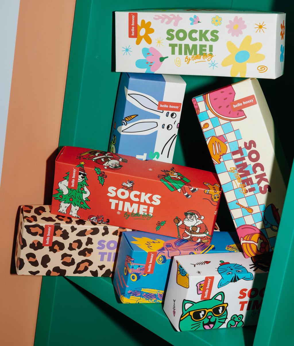 Coffrets cadeaux Socks Time ! empilés sur une étagère verte, packagings colorés et illustrés : motifs Noël, fleurs, chats, léopard, mascottes, voitures et nourriture – Hello Hossy